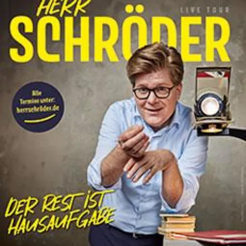 Ein fröhlicher Mann mit Brille steht an einem Tisch und hält eine alte Filmkamera. Der Text kündigt seine Live-Tour mit dem Titel "Der Rest ist Hausaufgabe" an.