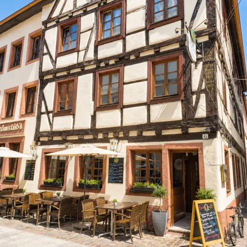 Ein charmantes Fachwerkhaus mit einer einladenden Terrasse. Tische und Stühle stehen draußen, umgeben von Pflanzen und Sonnenschirmen.