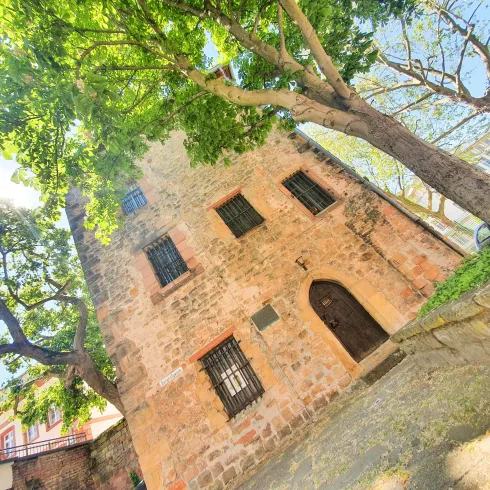 Ein historisches Gebäude aus Stein, umgeben von hohen Bäumen. Die Fassade hat kleine Fenster und eine Eingangstür.
