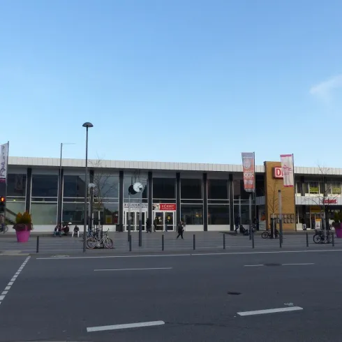 Ein modernes Einkaufszentrum mit großen Fenstern und bunten Blumenkübeln. Vor dem Gebäude stehen Fahrräder und es gibt mehrere Personen auf dem Gehweg.