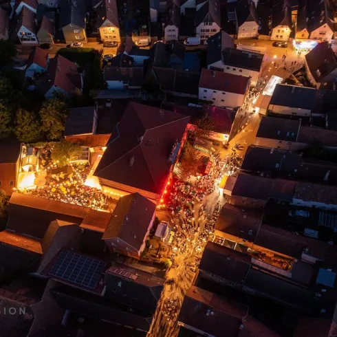 Eine belebte Stadt mit engen Gassen, erleuchtet von zahlreichen Lichtern. Menschen versammeln sich an einem zentralen Platz, umgeben von traditionellen Häusern.