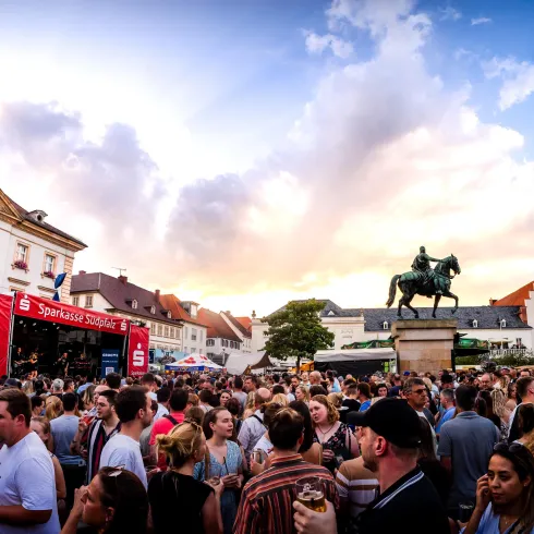 Ein belebter Platz mit einer großen Menschenmenge beim Fest. Im Hintergrund steht ein Denkmal und der Himmel ist teilweise bewölkt.