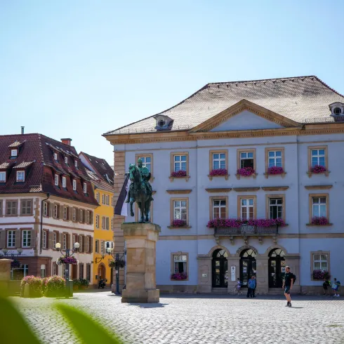 Ein schöner Platz mit einem historischen Gebäude und einem Denkmal. Umgeben von weiteren traditionellen Gebäuden und grünem Pflanzenbewuchs.