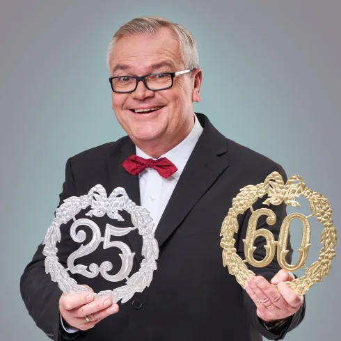 Ein fröhlicher Mann in einem Anzug hält zwei dekorative Kränze in den Händen. Einer ist silber und steht für 25 Jahre, der andere ist gold und symbolisiert 60 Jahre.