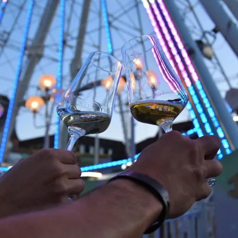 Zwei Gläser mit Wein werden hochgehalten, während im Hintergrund ein Riesenrad mit bunten Lichtern leuchtet. Die Atmosphäre ist feierlich und festlich.