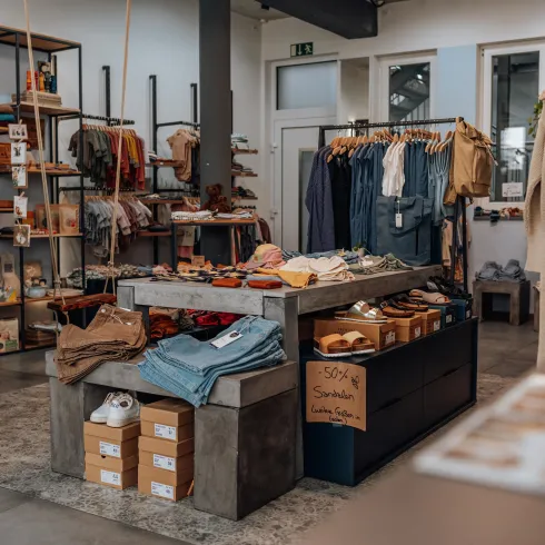 Ein gemütlicher Bekleidungsladen mit verschiedenen Kleidungsstücken und Accessoires. In der Mitte steht ein großer Tisch mit einer Auswahl an Schuhen und Jeans.