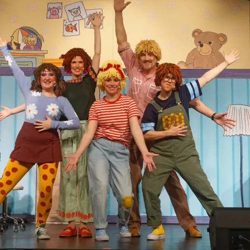 Eine Gruppe von fünf Personen in bunten Kostümen steht auf einer Bühne. Sie posieren fröhlich vor einer kindgerechten Kulisse.