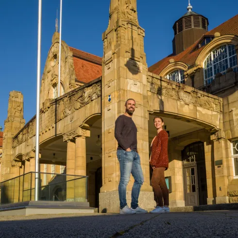 Zwei Personen stehen vor einem historischen Gebäude mit dekorativen Steinelementen. Der Himmel ist blau und die Sonne scheint, was eine freundliche Atmosphäre schafft.