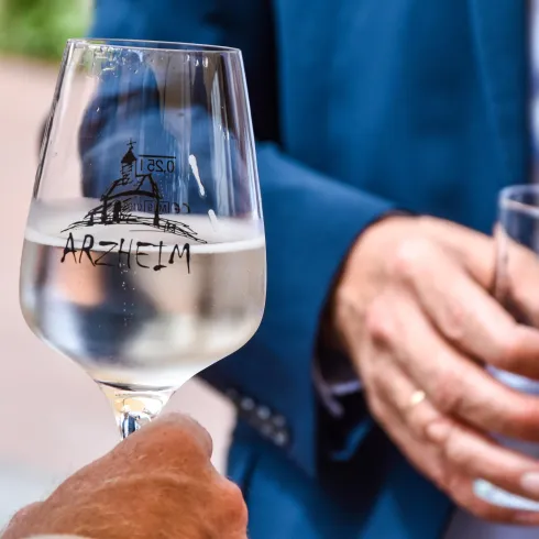 Ein Glas mit weißem Wein und der Aufschrift "Arztielm" wird gehalten. Im Hintergrund sind Hände mit einem Wasserglas zu sehen.