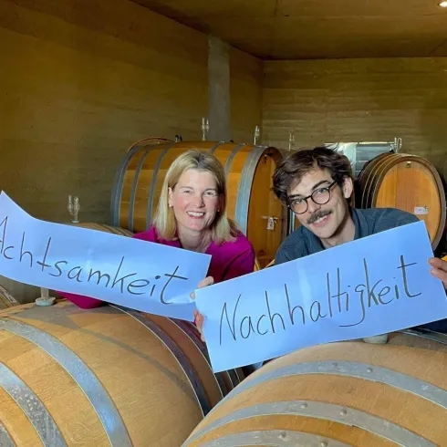 Zwei Personen stehen in einem Weinkeller und halten Plakate mit den Wörtern „Achtsamkeit“ und „Nachhaltigkeit“. Im Hintergrund sind Weinfässer zu sehen.