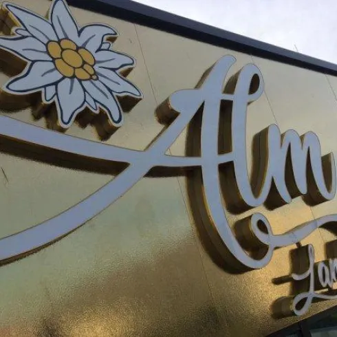 Ein goldenes Schild mit dem Schriftzug "Alm" und einer Edelweiss-Blume. Es zeigt eine elegante und einladende Präsentation.