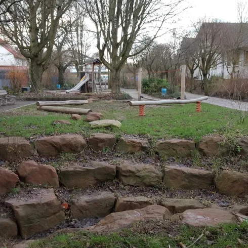 Ein Spielplatz mit Rasenflächen und Holzstämmen. Im Hintergrund sind Bäume und ein Spielgerät zu sehen.