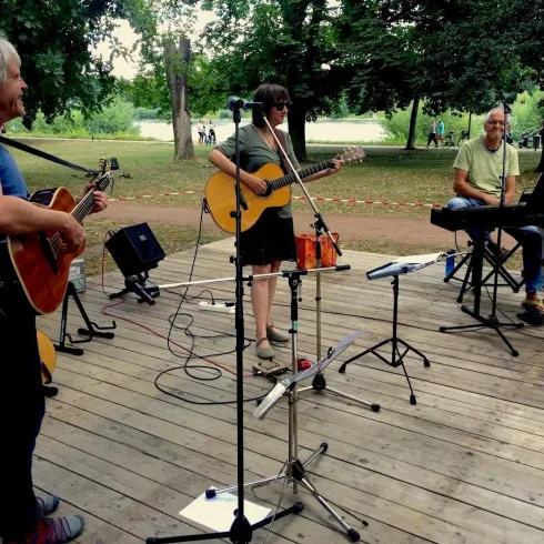 Eine Musikgruppe spielt im Freien auf einer Holzbühne. Drei Musiker mit Gitarren und einem Keyboard unterhalten das Publikum in einem Park.
