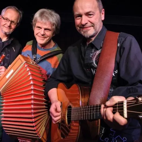 Drei Musiker stehen auf der Bühne und spielen verschiedene Instrumente. Einer hat eine Gitarre, während ein anderer ein Akkordeon hält.