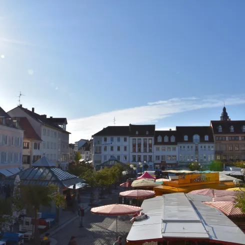 Ein lebhafter Marktplatz mit bunten Ständen und vielen Menschen. Im Hintergrund sind malerische alte Gebäude unter einem klaren blauen Himmel zu sehen.