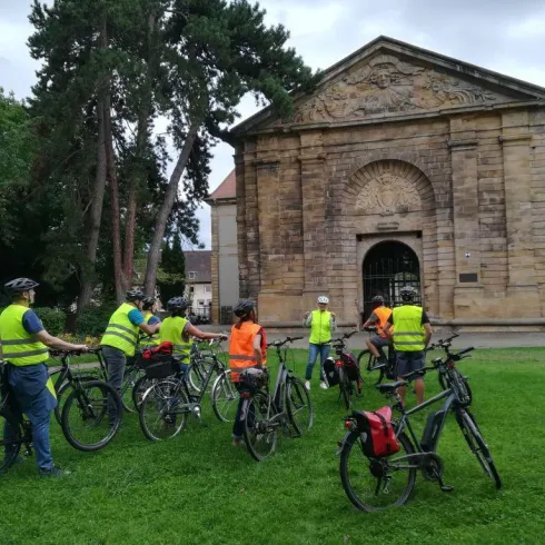 Eine Gruppe von Radfahrern in leuchtenden Westen steht auf einer Wiese vor einem historischen Gebäude. Im Hintergrund sind Bäume und ein bewölkter Himmel sichtbar.