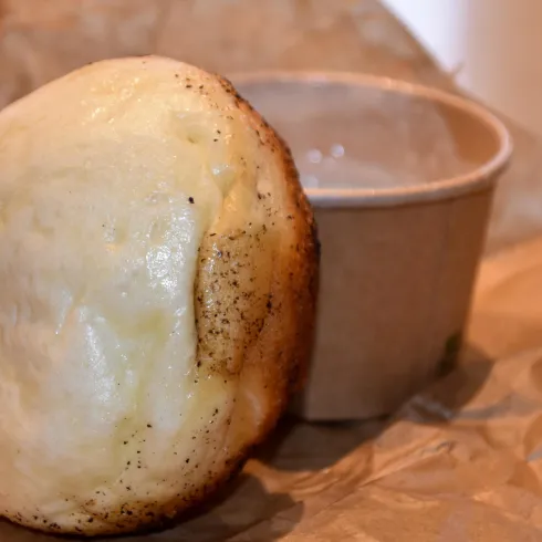 Ein frisch gebackenes Brötchen liegt neben einem kleinen Becher. Der Becher enthält wahrscheinlich eine Soße oder Dip.