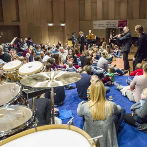 Eine Gruppe von Kindern und Erwachsenen sitzt auf dem Boden und hört einer musikalischen Darbietung zu. Im Vordergrund sind Schlaginstrumente zu sehen, während der Raum voller Musik und Freude ist.