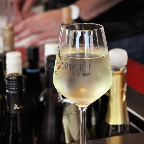 Ein Glas Weißwein steht vor einer Auswahl an Weinflaschen. Im Hintergrund ist eine Person beim Servieren von Getränken zu sehen.