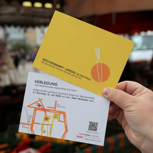 Eine Hand hält zwei Karten auf einem Markt. Die obere Karte ist gelb mit einem Karottenmotiv und die untere enthält eine Karte mit Wegbeschreibung.
