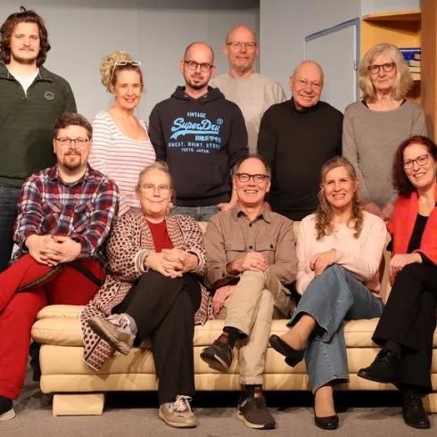 Eine Gruppe von Menschen sitzt auf einem Sofa und lächelt in die Kamera. Sie scheinen Teil eines Theater- oder Filmprojekts zu sein.