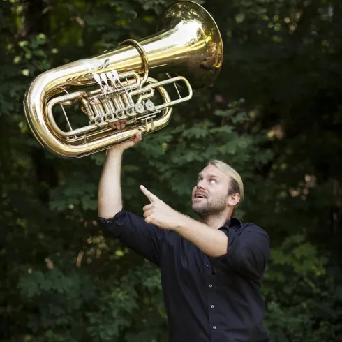 Ein Mann hebt eine große, glänzende Tuba in die Höhe. Er zeigt mit dem Finger darauf und lächelt.
