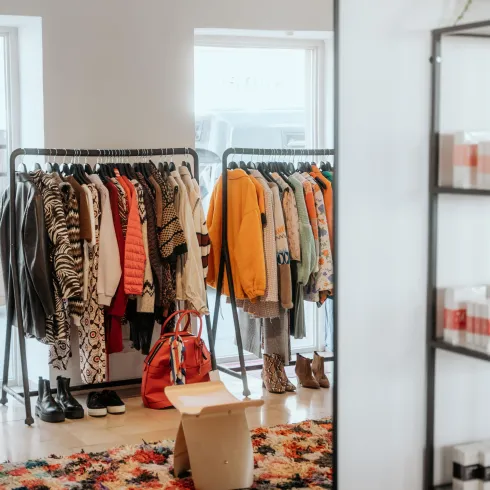 Ein modisches Geschäft mit einer Kleiderstange, die verschiedene Bekleidungsstücke zeigt. Im Hintergrund ist ein bunter Teppich und ein Regal mit Kosmetikprodukten zu sehen.
