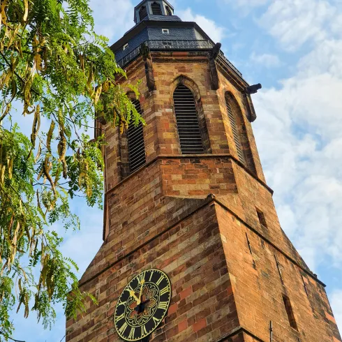 Ein historischer Turm mit einer großen Uhr und gotischen Fenstern. Der Himmel ist teilweise bewölkt und Bäume sind im Vordergrund sichtbar.