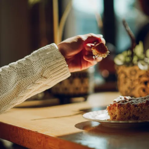 Eine Hand greift nach einem Stück Kuchen auf einem Teller. Im Hintergrund sind Pflanzen und eine weitere Person zu sehen.