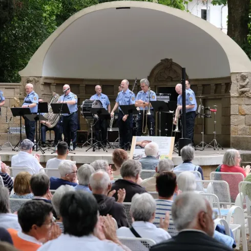 Eine Musikkapelle spielt im Freien vor einem Publikum. Die Zuschauer klatschen und genießen die Darbietung in einer schönen Umgebung.
