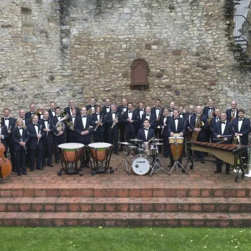Eine große Musikgruppe in Anzügen posiert vor einer alten Steinmauer. Sie spielen verschiedene Instrumente und stehen auf einer Stufe aus roten Steinen.
