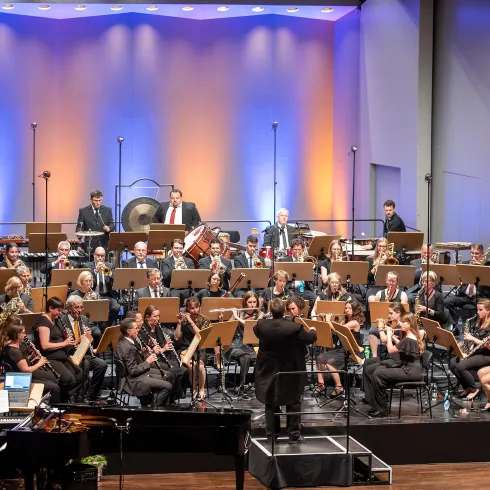 Ein Orchester spielt auf einer Bühne mit bunten Lichtern im Hintergrund. Der Dirigent leitet die Musiker, die verschiedene Instrumente spielen.