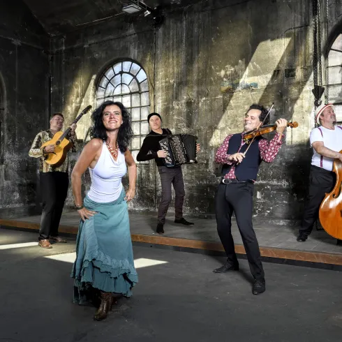 Vier Frauen in eleganten Kleidern sitzen um ein Klavier. Sie lächeln und posieren zusammen für das Foto.