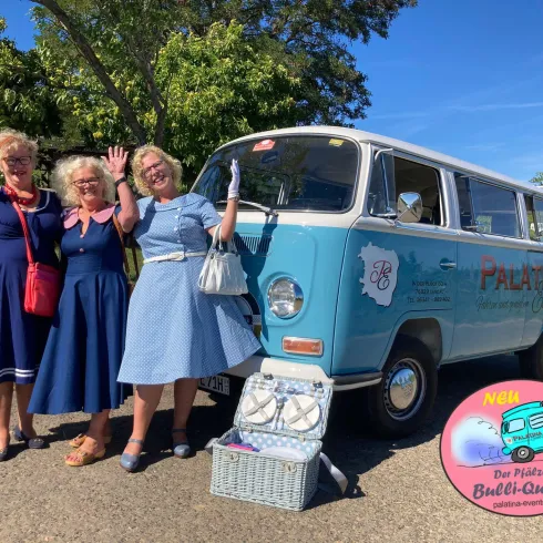 Drei Frauen in vintage Kleidern stehen fröhlich vor einem blauen VW-Bus. Im Hintergrund sind Bäume und ein klarer Himmel zu sehen.