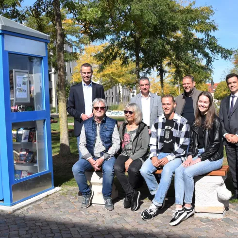 Eine Gruppe von Personen posiert vor einer blauen Telefonzelle. Der Hintergrund zeigt Bäume und einen sonnigen Tag.