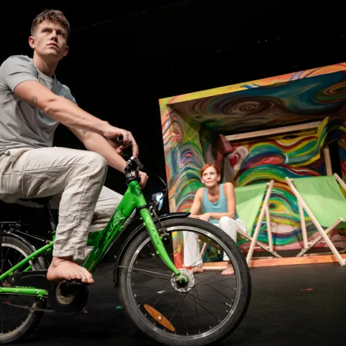 Ein junger Mann sitzt barfuß auf einem grünen Fahrrad, während eine Frau im Hintergrund auf einem Stuhl sitzt. Die Kulisse zeigt bunte kreative Elemente, die eine lebendige Szene schaffen.