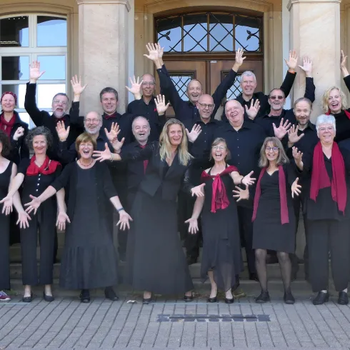 Eine fröhliche Gruppe von Sängern in schwarz-rot posiert vor einem Gebäude. Sie zeigen ihre Begeisterung mit erhobenen Händen und lächeln.