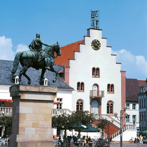 Ein beeindruckendes Reiterstandbild steht vor einem historischen Gebäude mit einer auffälligen Uhr. Die Szene ist lebhaft mit Menschen und Fahrrädern im Hintergrund.