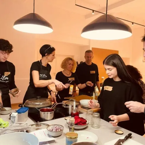 Eine Gruppe von Personen bereitet gemeinsam Essen in einer modernen Küche vor. Alle tragen schwarze Schürzen und arbeiten an verschiedenen Zutaten und Kochgeräten.