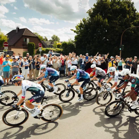 Eine Gruppe von Radfahrern fährt in einem Wettkampf vorbei. Zahlreiche Zuschauer stehen am Straßenrand und feuern sie an.