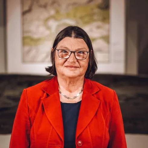 Eine Frau mit Brille trägt einen roten Blazer. Im Hintergrund sind Kunstwerke und eine ruhige Atmosphäre zu sehen.