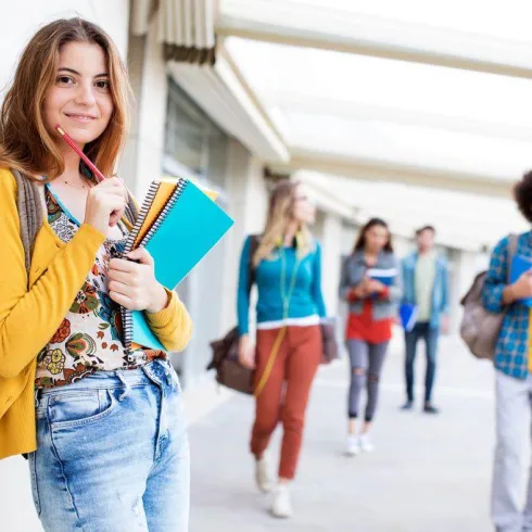 Ein fröhliches Mädchen mit Notizbuch und Stift lächelt an einer Wand. Im Hintergrund sind weitere Schüler, die sich unterhalten und im Flur gehen.