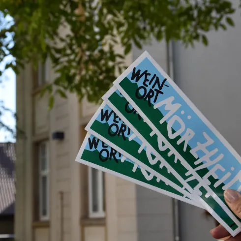 Eine Hand hält mehrere bunte Flyer mit dem Schriftzug "Mörlheim" und "Weinort". Im Hintergrund sind grüne Blätter und ein unscharfes Gebäude zu sehen.