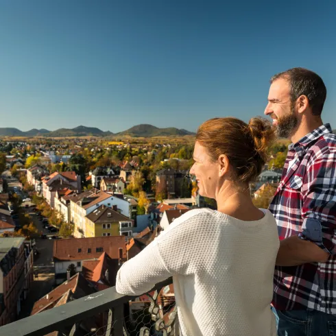 Ein Paar steht auf einem Aussichtspunkt und blickt auf die Stadt mit bunten Häusern und Hügeln im Hintergrund. Die Szene vermittelt eine entspannte Atmosphäre an einem sonnigen Tag.