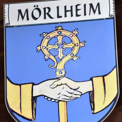 Ein Wappen von Mörlheim, das zwei Hände zeigt, die sich zur Begrüßung reichen. Darüber befindet sich ein goldenes Kreuz und der Name "Mörlheim".