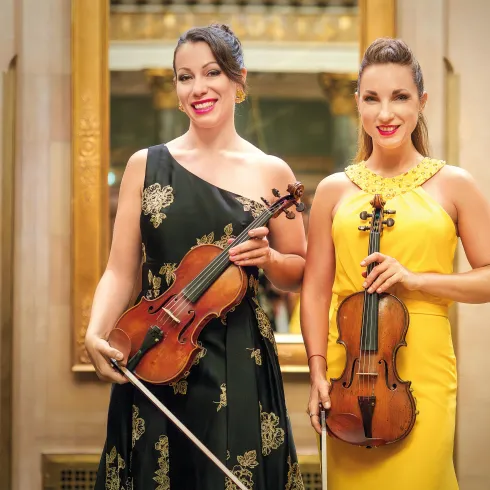 Zwei Musikerinnen stehen lächelnd in eleganter Kleidung. Sie halten Geigen und stehen vor einem geschmackvoll gestalteten Hintergrund.