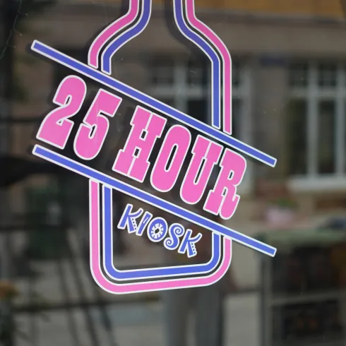 Ein bunter Aufkleber mit der Aufschrift "25 HOUR KIOSK". Der Hintergrund zeigt eine Stadtansicht mit Fenstern und Bäumen.