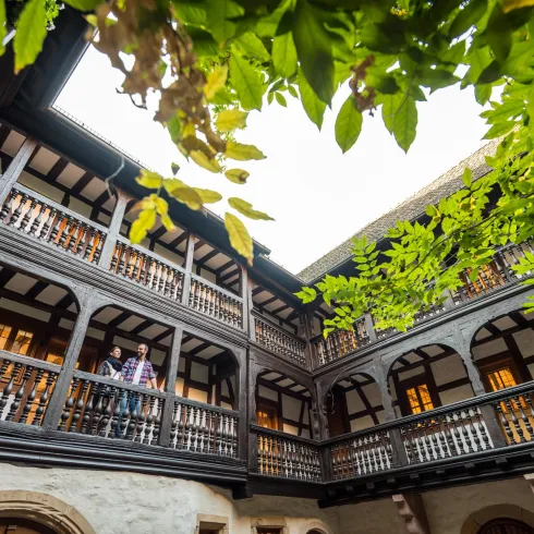 Ein historischer Innenhof mit Holzbalustraden und Pflanzen. Ein Mensch steht auf einem der Balkone und genießt die Atmosphäre.