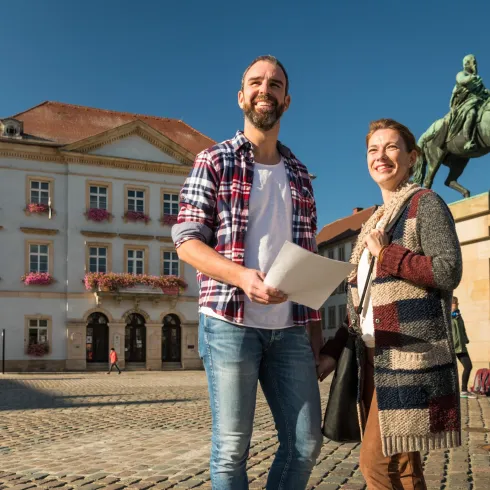 Ein sonniger Platz mit zwei lächelnden Personen, die vor einem historischen Gebäude stehen. Im Hintergrund sieht man ein Reiterstandbild und weitere Passanten.