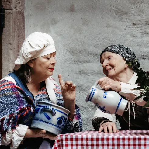 Zwei Frauen in traditioneller Kleidung unterhalten sich gemütlich an einem Tisch. Die eine hält einen Topf, während die andere mit einem Krug in der Hand zuschaut.